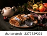 Homemade sliced traditional apple strudel pie in spotted ceramic plate served with ripe fresh apples, branches, sieve and icing sugar on dark linen tablecloth. Autumn mood tea drinking.