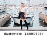 Young attractive blonde woman slung jacket over shoulder posing outdoors with sea and boats on background. Trendy girl in stylish outfit walking in a sea port.