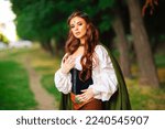 Small photo of Beautiful red haired princess in medieval elfin dress and long cloak with sword standing in warlike pose on green nature background. Fairy tale story about warrior in diadem . Warm colourful art work.