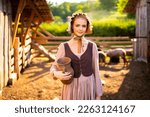 Beautiful young rural girl in medieval dress and bonnet hat posing on a ranch with jug of milk. Pretty blonde model in countryside art photo. 