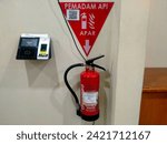 Small photo of Lampung, Indonesia - February 5th, 2024: A red fire extinguisher is hanging on the wall next to the timesheet machine for employees in a building at a private company.