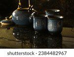Small photo of A dark setting with three blue teacups and matching blue teapot with lid. The teacups and teapot are on a dark countertop with water spilt beneath. The teacups have a brown and yellow rim.