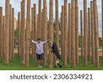 Small photo of A middle-aged couple plays between decorative wooden poles in a garden. He rests his hands on the poles, she hugs a pole.