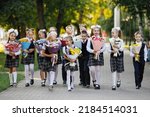 Small photo of Group of schoolchildren with flowers. Girls and boys in school uniforms walk along the sidewalk in a summer park. Selective focus, blurred background. Primary School.