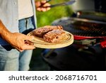 Small photo of Man put down on a wooden board ready to eat grilled steak meat, male roasted steak meat on the gas grill on barbecue grill outdoor in the backyard, summer family picnic, food on the nature.
