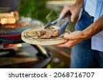 Small photo of Man put down on a wooden board ready to eat grilled steak meat, male roasted steak meat on the gas grill on barbecue grill outdoor in the backyard, summer family picnic, food on the nature.