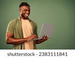 Small photo of Happy african american freelancer in casual khaki shirt looking on laptop screen while typing on keyboard. Positive handsome man standing over green background and surfing internet.