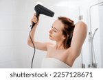 Young woman drying and styling hair with hair dryer, making hairstyle in modern bathroom interior. Beauty routine after morning shower