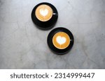 Two black mugs of cappuccino with heart shaped latte art on concrete background.