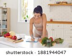 Small photo of Cheerful pretty slim female athete in sportswear standing in kitchen in sportswear and writing down healthy recipe or daily ration diet at home. Active healthy lifestyle, clean eating concept