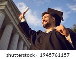 Small photo of Low angle portrait of happy triumphant male graduate standing near university holding up diploma. From below of young handsome man proud of academic achievements celebrating college graduation