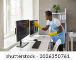 Small photo of Smiley caretaker, charwoman, daily janitor service lady in uniform with antiseptic spray bottle and wet cloth rag cleaning desktop PC computer, desk and table surfaces in modern office room interior