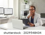 Small photo of Black woman reading documents and thinking sitting at office desk with modern laptop. Business lady in glasses considering question, studying contract terms and conditions, making important decision