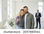 Small photo of Happy smiling young married couple looking at camera and showing key to new home standing in unfurnished white living room with real estate agent or mortgage broker in background. Buying house concept