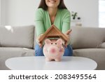Small photo of Woman saving up money to buy her own house. Happy young girl holding roof above small pink piggy bank placed on table in living room, close up. Finance, property purchase, taking mortgage loan concept