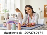 Small photo of Portrait of woman dressmaker, fashion designer, tailor or seamstress at work in studio. Smiling woman looking at camera while working with fabric while sitting at her workplace in bright sewing studio