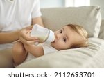 Small photo of Baby enjoying nutritious meal. Mother feeding baby on sofa. Nanny formula feeding little child as alternative to breastfeeding. Baby boy or girl lying on couch and drinking milk from plastic bottle