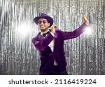 Small photo of Happy goofy young man wearing a ridiculous tacky unfashionable purple velvet jacket and hat doing funny dance moves at a party. Hilarious dorky guy dancing to music on a silver foil fringe background