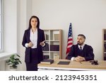 Small photo of Lawyer or defense attorney making speech during trial session in court. Serious unsmiling woman who represents defendant in lawsuit or criminal prosecution speaking to judge and audience in courtroom