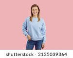Young model posing in studio. Studio shot of cheerful woman in casual outfit. Happy smiling teen girl wearing pale blue sweatshirt and jeans standing isolated on solid pastel pink colour background
