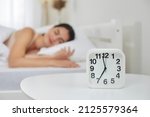 Alarm clock that is going to ring at seven in morning on background of woman sleeping in bed. Close up of alarm clock standing on table next to young woman sleeping sweetly in her bed. Selective focus