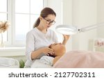 Small photo of Female cosmetologist performs facial examination procedure at cosmetology clinic. Beautician fixes lamp over face of female patient lying on examination couch in beauty clinic. Cosmetology concept.