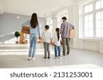 Small photo of Family buying house or apartment and moving to new home. Back view mom, dad and kids holding cardboard boxes standing in beautiful empty unfurnished spacious living room interior with big white window