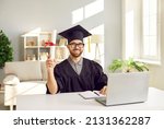 Small photo of Portrait of happy nerdy student in graduate mortarboard hat and robe sitting at desk with laptop, holding diploma scroll, looking at camera and smiling. University education and online degree concept