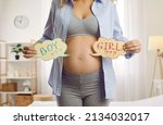 Small photo of Expectant mom trying to guess what gender her baby will be. Happy young pregnant woman with a big bare belly standing in the bedroom at home and holding two cute cards that read BOY and GIRL