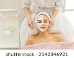 Small photo of Satisfied young Caucasian beautiful woman uses services of professional beautician in beauty salon or spa. Unrecognizable beautician applied white mask to half of client's face with brush. Top view.