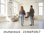 Small photo of This is our home now. Young married couple settling down in new place. Rear view from behind of two people who are moving house or apartment standing in spacious room, holding boxes and looking around