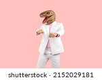Small photo of Weird guy in funny disguise dancing against pastel pink studio background. Cheerful eccentric man in white suit and silly ugly wacky masquerade dinosaur mask having fun at crazy party