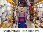 Happy salesman at modern DIY store guarantees best quality of all the goods. Portrait of handsome young man in uniform standing between shelves with ventilation grids, smiling and showing thumbs up