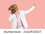 Small photo of Funny weird guy in wacky animal mask having fun at crazy party. Eccentric man in white suit and silly ugly masquerade dinosaur mask dancing isolated on pink background