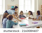 Small photo of Preschool teacher helps a group of children put together a puzzle. A woman teacher helps children in a preschool group. A group of children at the table collecting a puzzle