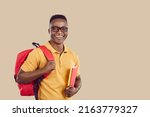 Happy African American school, college or university student with backpack. Cheerful handsome young man in polo shirt and glasses standing on beige copy space background, holding books and smiling