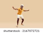 Small photo of Cheerful stylish african american man having fun dancing on light beige background. Funny smiling dark skinned man in sneakers, shorts, polo shirt and hat with sunglasses. Full length. Banner.