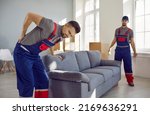 Small photo of Worker feels sudden pain in his lower back when he tries to lift a heavy sofa. Two young men from a delivery service or a moving company can't carry a heavy couch because one of them gets injured