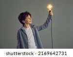 Small photo of Happy little boy holding shining lit Edison light bulb standing on gray studio background. Smart kid looking at glowing incandescent lightbulb in his hand. Creative idea and inspiration concept