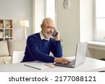 Small photo of Happy retired senior man talking on his mobile phone. Cheerful bald man in glasses sitting at his working table, speaking to a friend on his cellphone and using his laptop at the same time