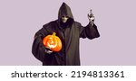 Small photo of Man in image of mystical creepy grim reaper with pumpkin in his hand raises finger in warning with idea. Man with skull make-up and in long black cloak with hood looks down on light lilac background
