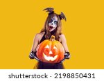 Small photo of Portrait of child in creative Halloween costume. Girl with skull makeup standing isolated on yellow background, holding Jack o lantern pumpkin and looking at camera with surprised face expression