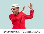 Young energetic African American man wearing sunglasses and red entertainment suit, yelling and clapping hands performing funny dance or working as host of party stands on pastel blue background