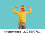 Small photo of Man in funny chicken disguise pretends to be strong and have superpower. Overconfident young guy wearing strange ridiculous bizarre rooster mask flexing his weak arms isolated on turquoise background