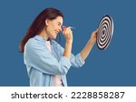 Small photo of Happy beautiful confident young brunette woman in casual shirt squints eye and aims dart arrow at shooting target in her hand. Side profile view studio shot. Setting goals, strategy, success concept