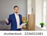 Small photo of Portrait of professional realtor or real estate agent. Happy young man in suit standing in living room of new house for sale, holding clipboard, looking at camera and smiling