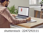 Small photo of Concentrated male financier using laptop working in office with data entered in spreadsheet. Man fills out paper documents and enters data into electronic files marked in red on laptop screen.