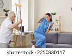 Small photo of Angry aggressive elderly man threatening to his caregiver woman. Elderly patient suffering from mental disability threatening with crutch to frightened nurse. Professional medical help and support