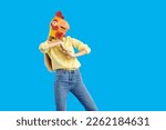 Small photo of Woman in funny strange crazy disguise dancing isolated on solid blue background. Studio portrait of slim young model wearing casual shirt, jeans and wacky goofy yellow chicken, hen or rooster mask