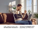 Smiling young man with broken arm making video call using laptop. Cheerful handsome man wearing arm splint sitting on sofa at home and waving his hand in greeting having meeting online